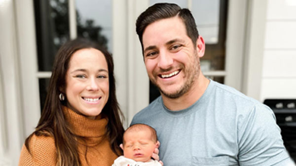 A smiling couple holding a newborn baby stands in front of a house with large windows. the woman is wearing a brown turtleneck and the man is in a gray t-shirt.