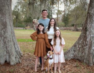 A smiling family of four, two adults and two young girls, pose outdoors between large trees with two dogs. The girls wear dresses; the adults stand behind them, one holding a small dog. The background shows grass and trees.