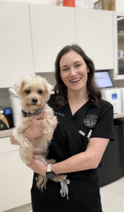 A smiling woman in black scrubs holds a small, fluffy dog in a veterinary clinic. Veterinary equipment and supplies are visible in the background.