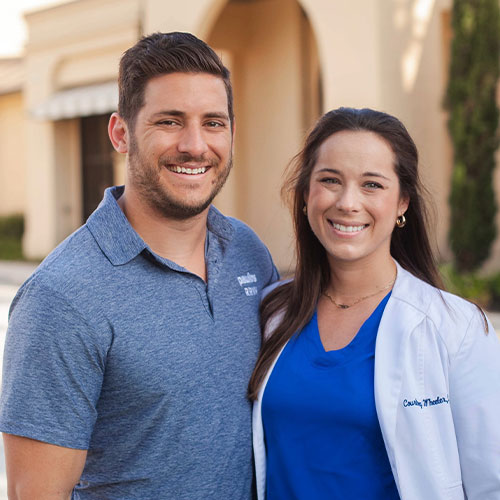 A smiling man and woman standing outdoors. the man wears a casual grey polo shirt while the woman wears a blue medical scrubs top and a white lab coat.