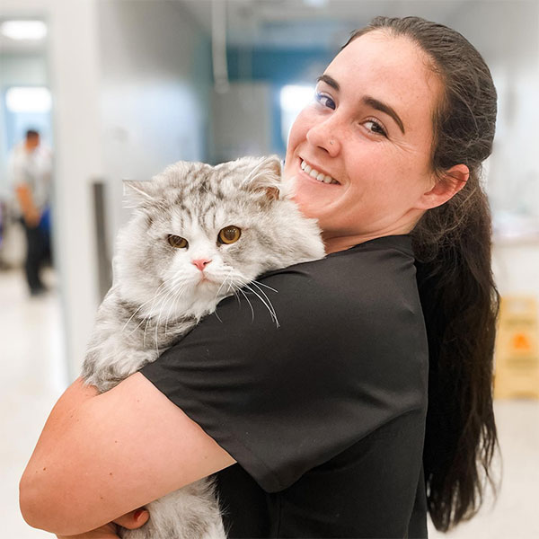 A woman smiling warmly as she holds a fluffy gray cat in her arms. the background is softly blurred, indicating an indoor setting.
